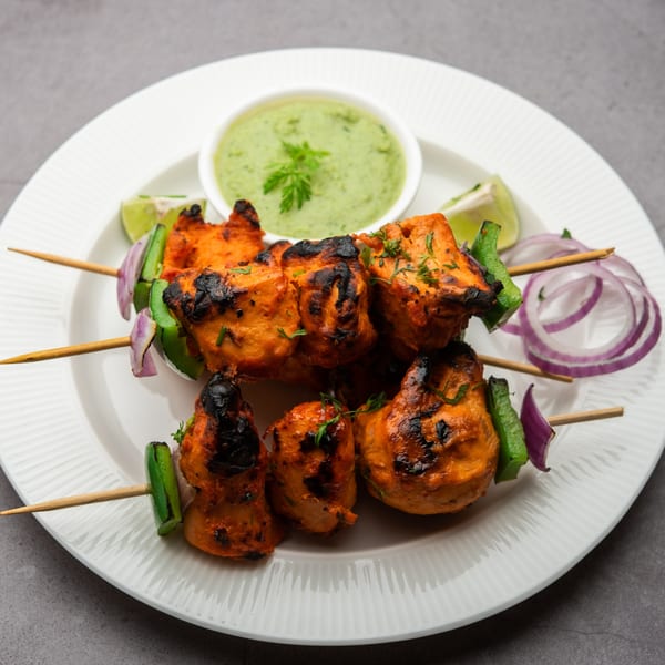Grilled chicken tikka skewers with green chutney, lime, and red onion on a white plate.